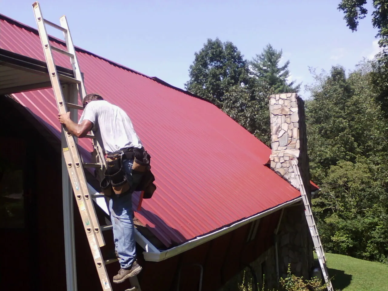 Roofer on a ladder beside a red metal roof and stone chimney — Soffit & Fascia Repair by Roofing Brothers in Preston